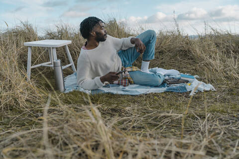 Young man with smart phone looking away while lying on blanket amidst dried plants stock photo