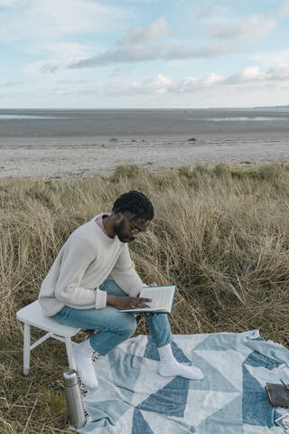 Junger Mann liest ein Buch und sitzt auf einem Hocker am Strand, lizenzfreies Stockfoto