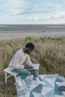 Junger Mann liest ein Buch und sitzt auf einem Hocker am Strand - BOYF01875