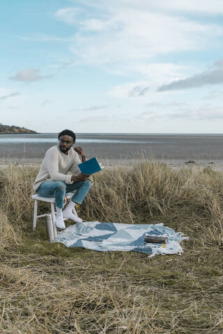 Junger Mann mit Buch, der wegschaut, während er auf einem Hocker am Strand vor einem bewölkten Himmel sitzt, lizenzfreies Stockfoto