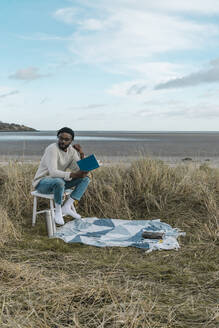 Junger Mann mit Buch, der wegschaut, während er auf einem Hocker am Strand vor einem bewölkten Himmel sitzt - BOYF01876