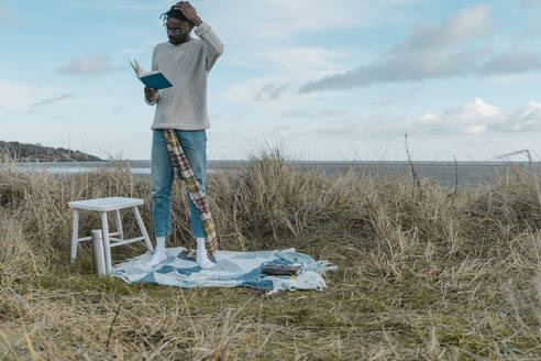 Junger Mann liest ein Buch, während er inmitten von getrockneten Pflanzen am Strand steht - BOYF01877