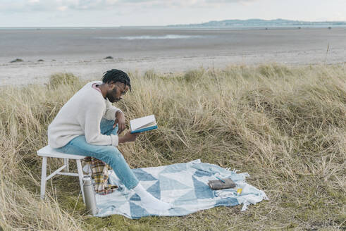 Konzentrierter junger Mann liest ein Buch, während er auf einem Hocker inmitten von getrockneten Pflanzen am Strand sitzt - BOYF01880