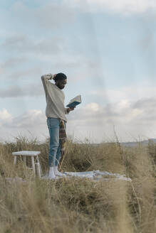 Junger Mann mit Hand im Haar, der ein Buch liest, während er vor einem bewölkten Himmel steht - BOYF01881