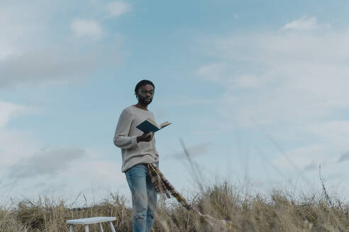 Man with book standing amidst dried plants at beach against cloudy sky - BOYF01882