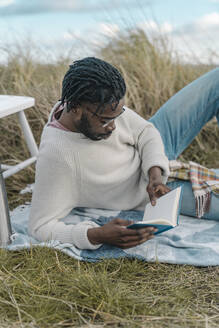 Entspannter junger Mann, der inmitten von getrockneten Pflanzen liegt und ein Buch liest - BOYF01883