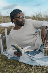 Smiling young man lying on blanket with book looking away - BOYF01884