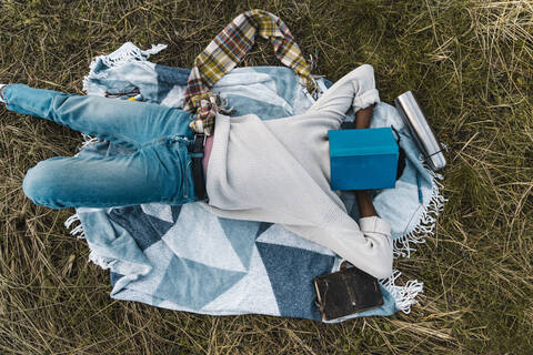 Junger Mann schläft auf einer Decke und bedeckt sein Gesicht mit einem Buch inmitten von getrockneten Pflanzen, lizenzfreies Stockfoto