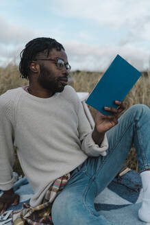 Junger Mann mit Brille liest ein Buch im Sitzen vor bewölktem Himmel - BOYF01888