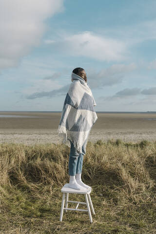 Junger Mann in eine Decke eingewickelt auf einem Hocker am Strand gegen den bewölkten Himmel stehend, lizenzfreies Stockfoto