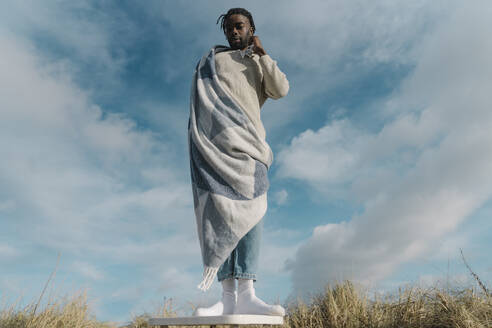 Junger Mann in Decke eingewickelt auf Hocker stehend gegen bewölkten Himmel - BOYF01898
