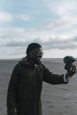 Smiling male vlogger live streaming through camera at beach against sky stock photo