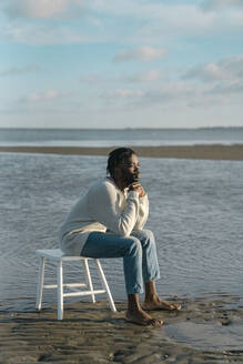 Young man sitting on white stool while watching sunset at beach - BOYF01906