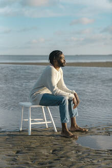 Young man sitting on white tool while watching sunset at beach - BOYF01907