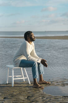 Entspannter junger Mann mit geschlossenen Augen sitzt auf weißem Hocker am Strand gegen den Himmel - BOYF01908