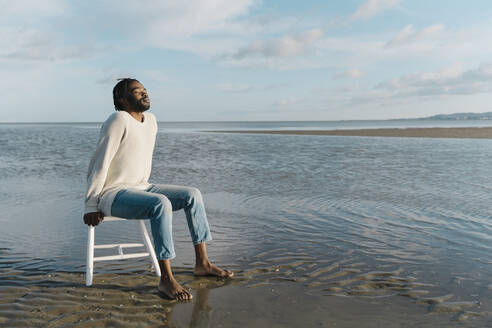 Thoughtful young man with eyes closed sitting on white stool at beach during sunset - BOYF01910