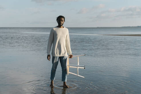 Junger Mann mit weißem Hocker steht am Strand gegen den Himmel - BOYF01916