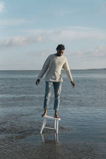 Young man standing on white stool while balancing at beach against cloudy sky - BOYF01917