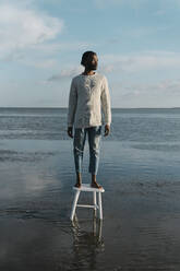 Nachdenklicher junger Mann, der bei Sonnenuntergang auf einem Hocker am Strand steht und wegschaut - BOYF01918