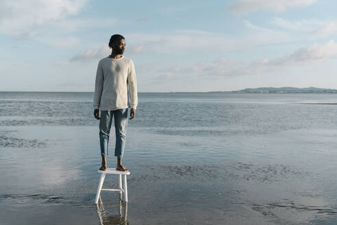 Thoughtful young man looking away while standing on stool at beach during sunset - BOYF01919