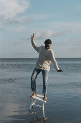 Junger Mann balanciert auf weißem Hocker am Strand gegen bewölkten Himmel - BOYF01920