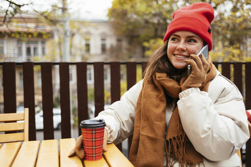 Frau in warmer Kleidung, die in einem Straßencafé sitzt und mit ihrem Handy telefoniert - OYF00321