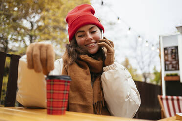 Lächelnde Frau, die einen Einwegbecher betrachtet, während sie in einem Straßencafé mit ihrem Mobiltelefon telefoniert - OYF00324