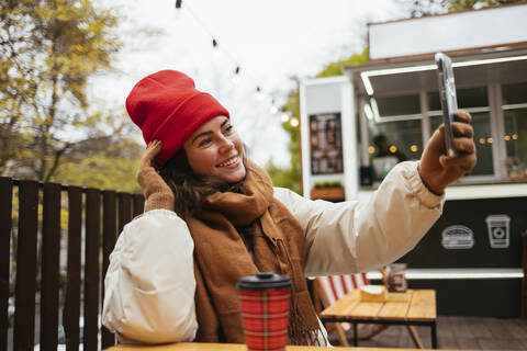 Frau in warmer Kleidung, die ein Selfie mit ihrem Mobiltelefon macht, während sie in einem Straßencafé sitzt, lizenzfreies Stockfoto