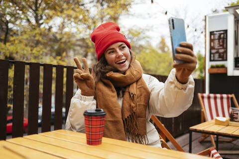 Happy woman gesturing peace sign while taking selfie through mobile phone sitting at sidewalk cafe stock photo