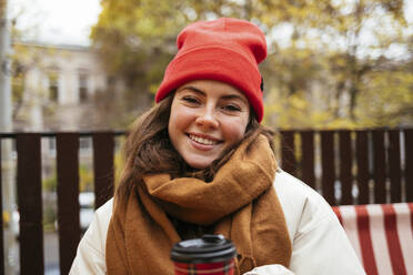 Junge Frau mit Strickmütze und Schal, die lächelnd in einem Straßencafé sitzt - OYF00329