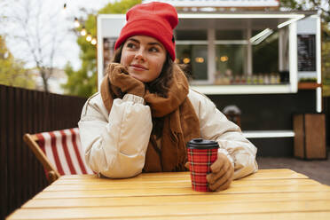 Frau in warmer Kleidung schaut weg, während sie mit der Hand am Kinn in einem Straßencafé sitzt - OYF00330