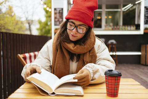 Frau mit Brille, die in einem Straßencafé sitzt und ein Buch liest, lizenzfreies Stockfoto