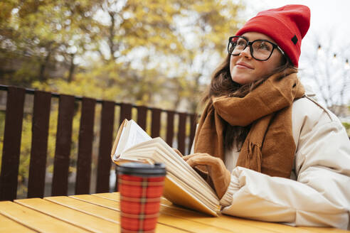 Lächelnde Frau mit Buch, die wegschaut, während sie in einem Straßencafé sitzt - OYF00333