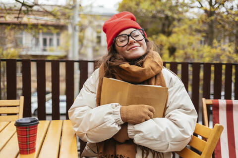 Frau in warmer Kleidung, die ein Buch umarmt, während sie in einem Cafe sitzt, lizenzfreies Stockfoto