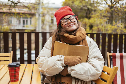 Frau in warmer Kleidung, die ein Buch umarmt, während sie in einem Cafe sitzt - OYF00336