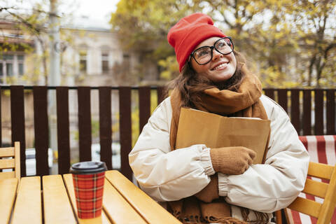 Nachdenkliche Frau lächelt, während sie ein Buch umarmt und in einem Straßencafé sitzt, lizenzfreies Stockfoto