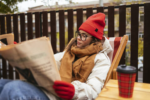 Junge Frau in warmer Kleidung liest Zeitung und entspannt sich auf einem Stuhl in einem Café, lizenzfreies Stockfoto