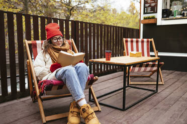Junge Frau in warmer Kleidung liest ein Buch und sitzt auf einem Stuhl in einem Straßencafé - OYF00341