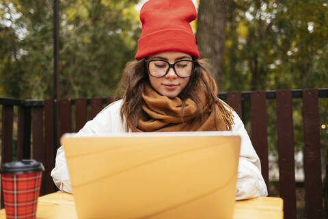 Frau mit Strickmütze und Schal, die in einem Straßencafé sitzt und einen Laptop benutzt, lizenzfreies Stockfoto