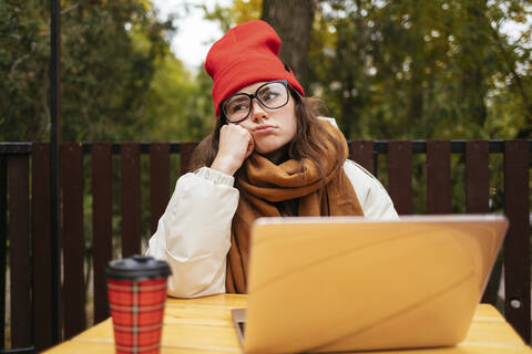 Gelangweilte Frau, die mit ihrem Laptop in einem Straßencafé sitzt und wegschaut, lizenzfreies Stockfoto