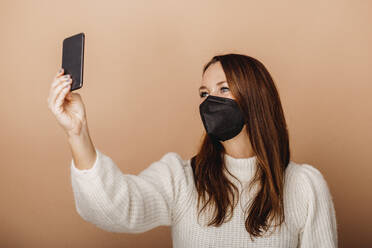 Junge Frau mit Gesichtsschutzmaske, die ein Selfie mit ihrem Mobiltelefon macht, während sie vor einem beigen Hintergrund steht - DAWF01758