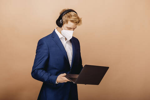 Geschäftsmann im Anzug und mit Gesichtsschutzmaske, der einen Laptop benutzt, während er vor einem beigen Hintergrund steht, lizenzfreies Stockfoto