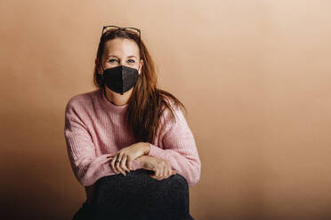 Young woman wearing protective face mask staring while standing against beige background - DAWF01762