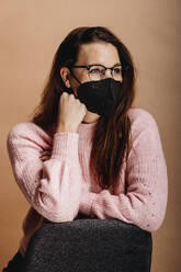 Woman wearing face mask looking away while standing by chair against beige background - DAWF01763