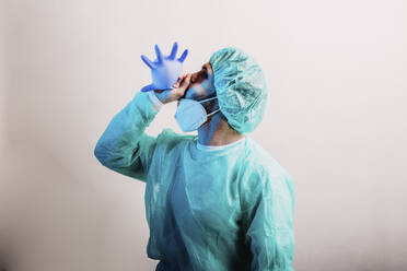Männlicher Arzt im Schutzanzug, der chirurgische Handschuhe bläst, während er vor einem grauen Hintergrund steht - DAWF01764
