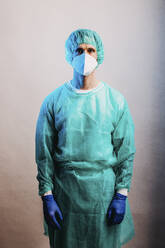Healthcare worker in protective workwear and face mask staring while standing against gray background - DAWF01765