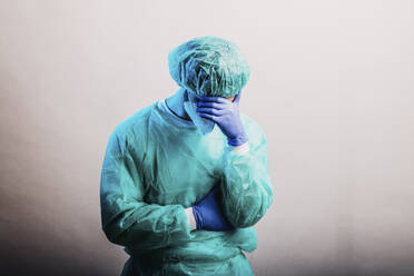 Male healthcare worker in protective suit and face mask standing with hand in hands against gray background - DAWF01766