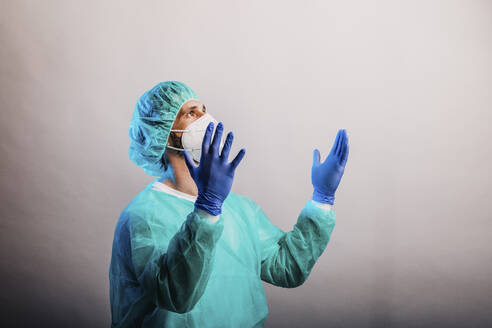 Frustrierter Arzt in Arbeitsschutzkleidung, der wegschaut, während er mit erhobener Hand vor grauem Hintergrund steht - DAWF01767