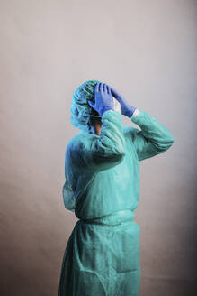 Männlicher Arzt in Schutzanzug, stehend mit dem Kopf in den Händen vor grauem Hintergrund - DAWF01768