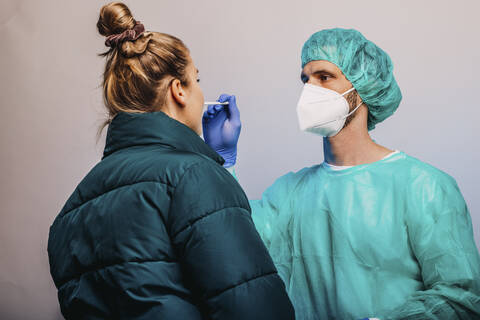 Männlicher Fachmann, der den medizinischen Test COVID-19 eines Patienten durchführt, während er vor einem grauen Hintergrund steht, lizenzfreies Stockfoto